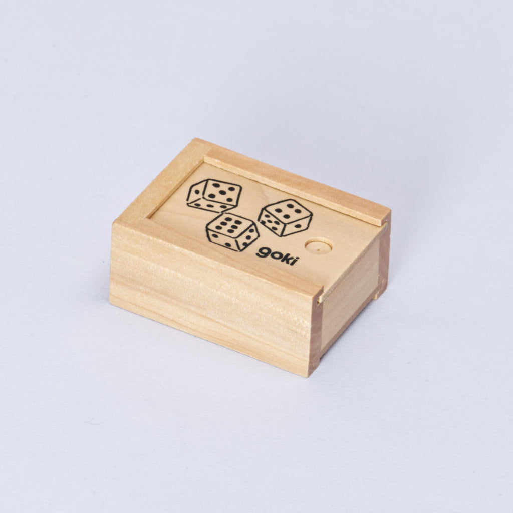 Eine Schachtel aus Holz mit Schiebedeckel, die sechs Würfel beinhaltet von goki ist seitlich von oben zu sehen. Der Deckel ist geschlossen und trägt die Aufschrift goki unten rechts und drei gezeichnete Würfel mit der Augenanzahl fünf, sechs und vier.