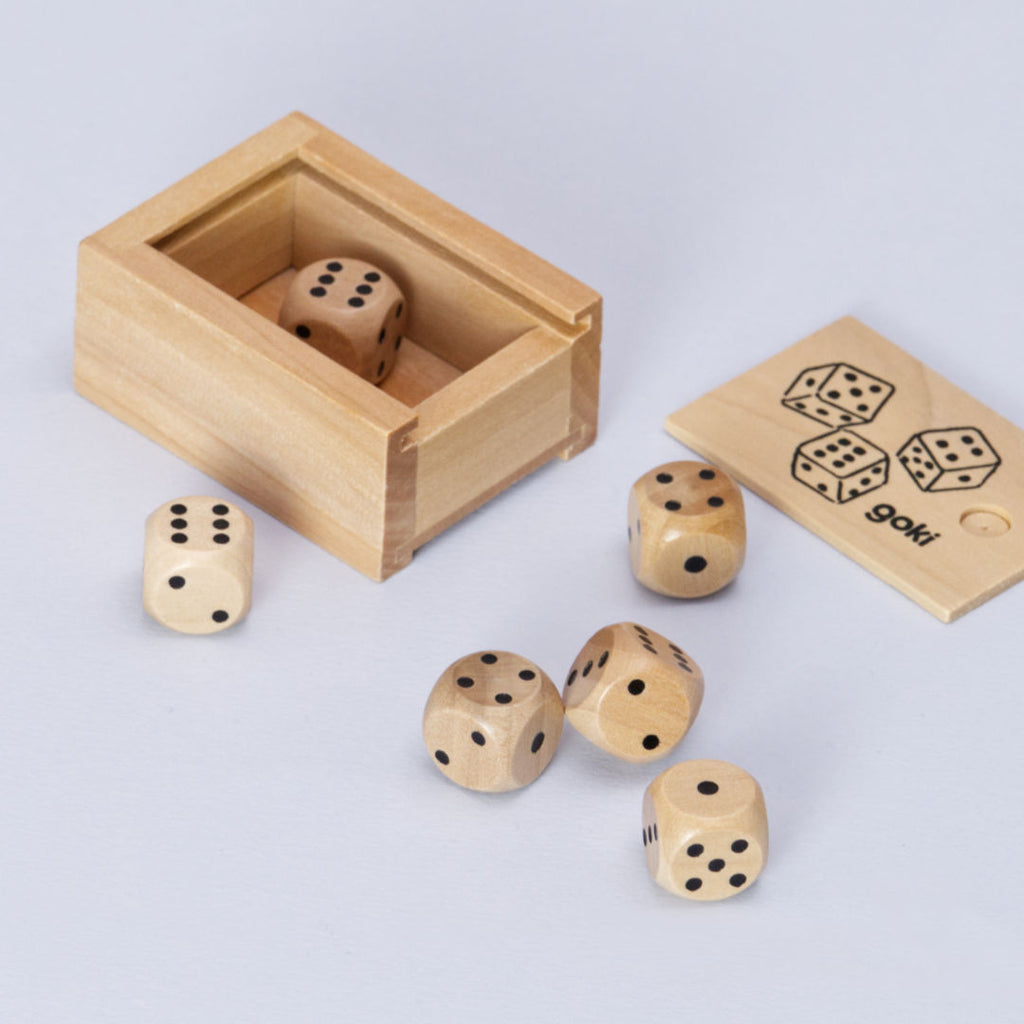 Eine Kiste aus Holz mit sechs Würfel von goki ist seitlich von oben zu sehen. Der Deckel liegt von der Kiste entfernt und trägt die Aufschrift goki unten rechts und drei gezeichnete Würfel. Einer der Würfel ist in der Schachtel, die anderen fünf liegen außerhalb.
