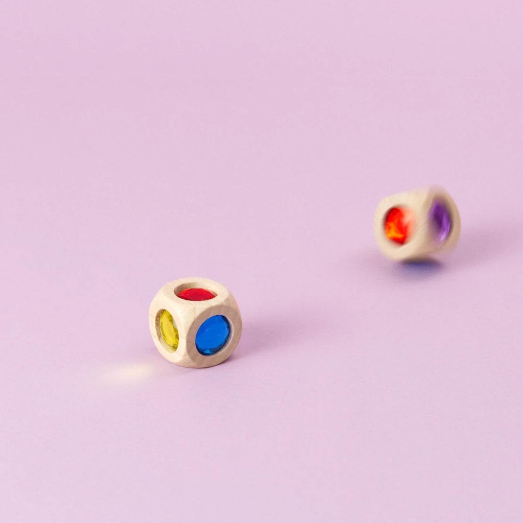 Ein Würfel aus Buchenholz mit funkelnden Acrylglas-Diamanten in Bewegung, unscharf geworfen, daneben ein liegender Würfel. Die Farben der Würfelseiten sind Rot, Blau, Grün, Gelb, Orange und Violett sichtbar.