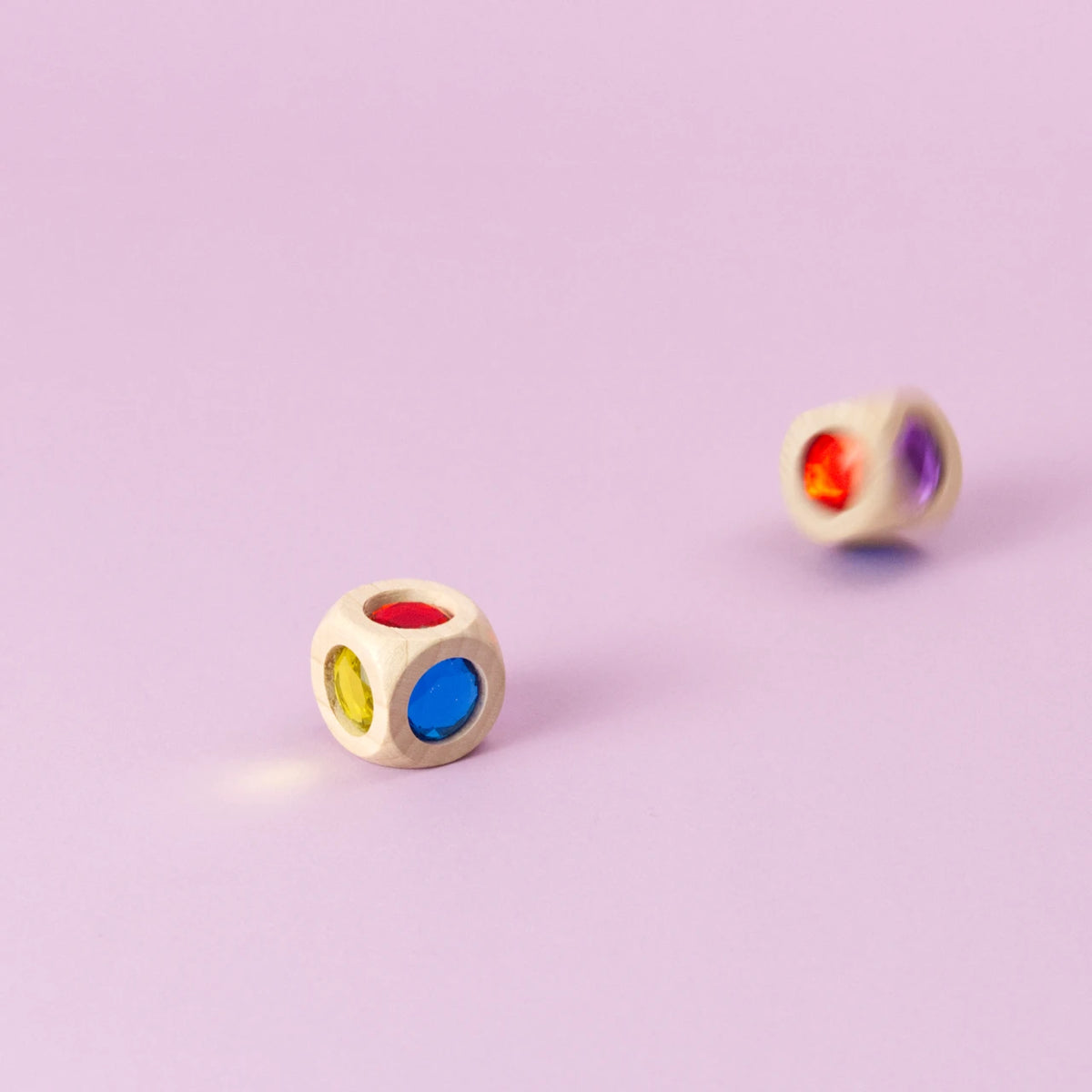Ein Würfel aus Buchenholz mit funkelnden Acrylglas-Diamanten in Bewegung, unscharf geworfen, daneben ein liegender Würfel. Die Farben der Würfelseiten sind Rot, Blau, Grün, Gelb, Orange und Violett sichtbar.