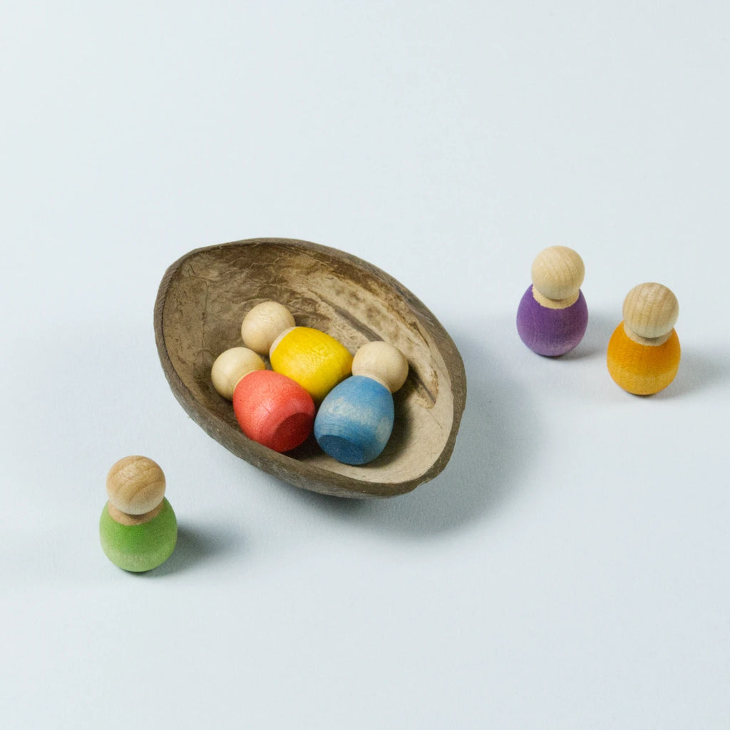 sechs kleine Babyholzfiguren in verschiedenen Farben in einer halben Kokosnussschale liegend und daneben stehend.