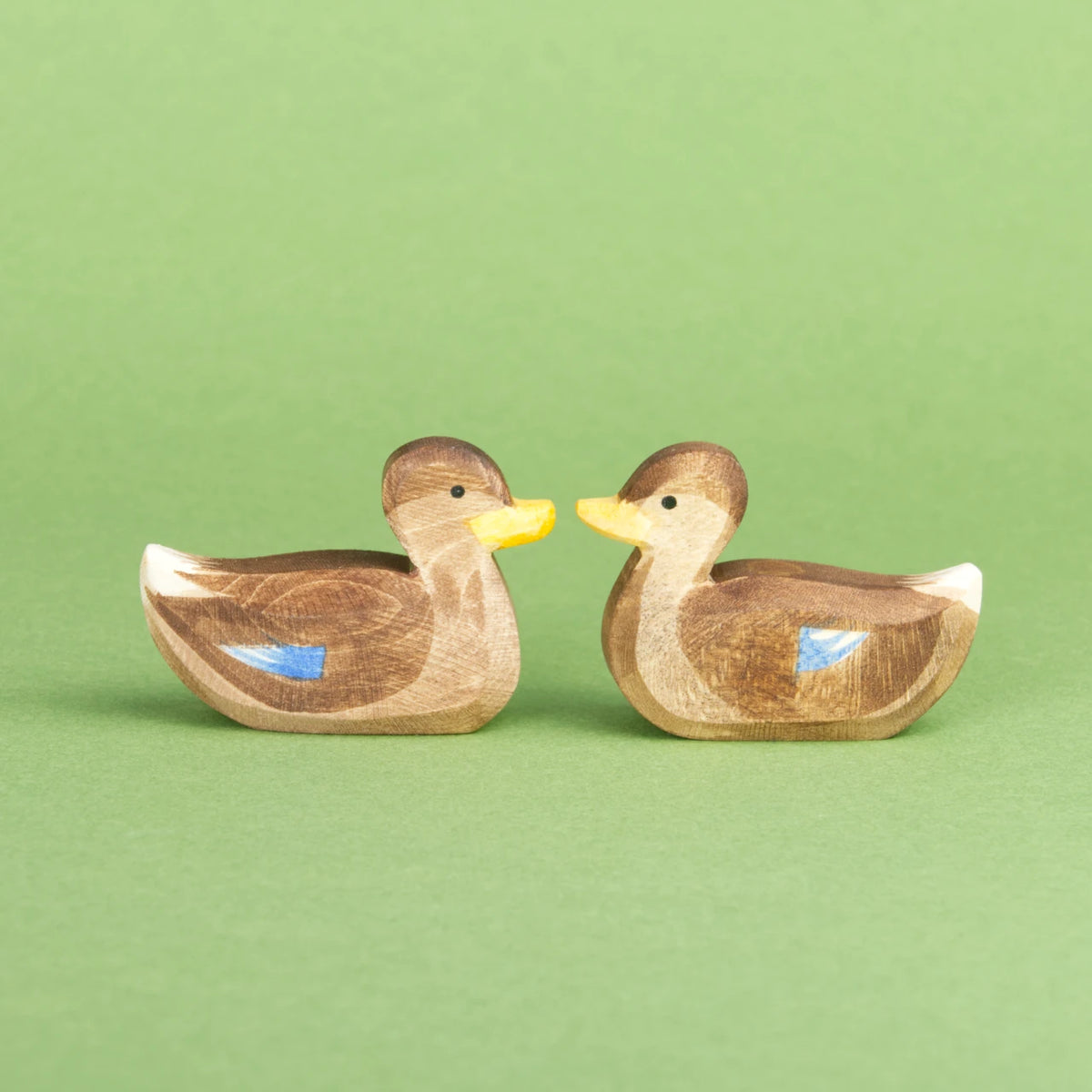 Zwei schwimmende Enten von Ostheimer aus Naturholz gefertigt, haben braunes Gefieder mit blauen und weißen Farbdetails, einen orangenen Schnabel und ein weißes Schwanzende. Beide schauen sich gegenseitig zur Bildmitte hin an.