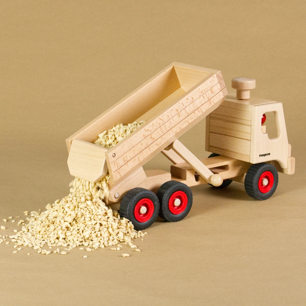 Muldenkipper-LKW von fagus aus naturlackiertem Buchenholz mit Vollgummireifen. Die Ladefläche ist hochgekippt, aus der Ladeklappe wird das Schüttgut entladen.
