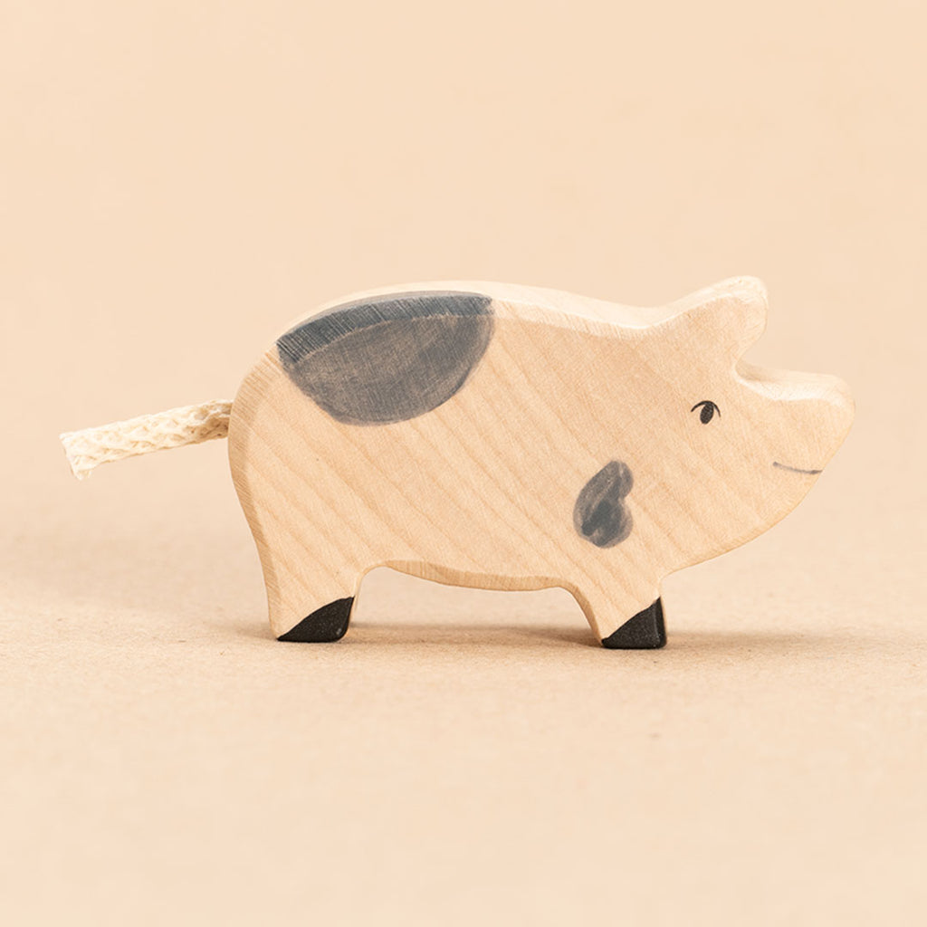 Ein aus Ahorn- und Buchenholz gefertigtes Ferkel, im Profil nach rechts schauend. Mit zwei grauen, organischen Flecken, sowie einem beigen Schwanz aus einem Stück Kordel. Nahaufnahme.