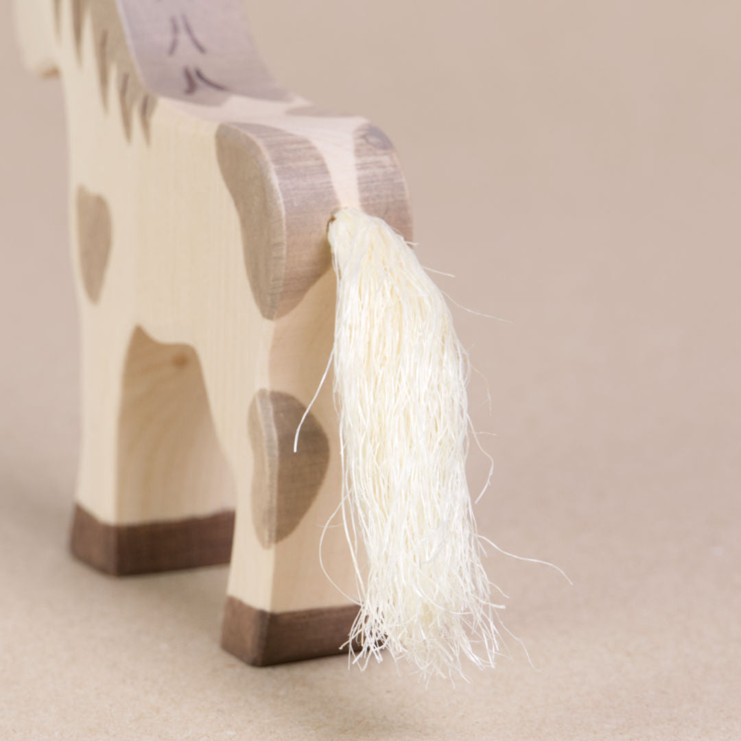 Hinteransicht eines nach rechts schauenden gefleckten Fohlens von Holztiger, es hat weiß, grau geflecktes Fell und einen Schweif aus Naturhaar. Die Hufen sind braun.