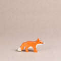 Ein nach rechts schauender, kleiner Fuchs von Holztiger, ist orange und hat eine weiße Schwanzspitze und weißes Bauchfell. Die Ohren stehen gerade nach oben.