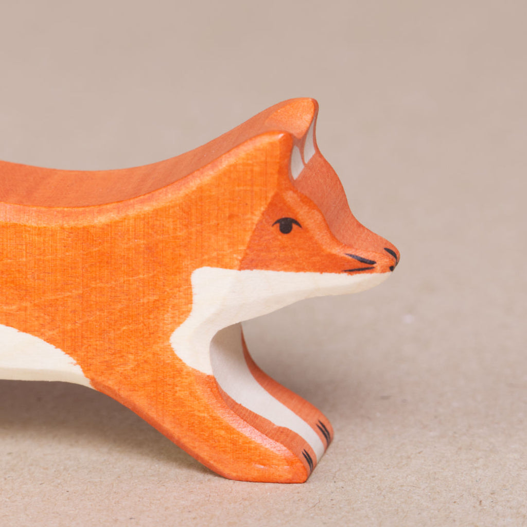 Nahaufnahme eines nach rechts schauenden, grossen, laufenden Fuchses von Holztiger, es ist orange und hat eine weiße Schwanzspitze und weißes Bauchfell. Die Ohren stehen gerade nach oben.
