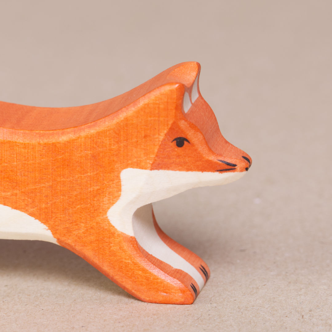 Nahaufnahme eines nach rechts schauenden, grossen, laufenden Fuchses von Holztiger, es ist orange und hat eine weiße Schwanzspitze und weißes Bauchfell. Die Ohren stehen gerade nach oben.