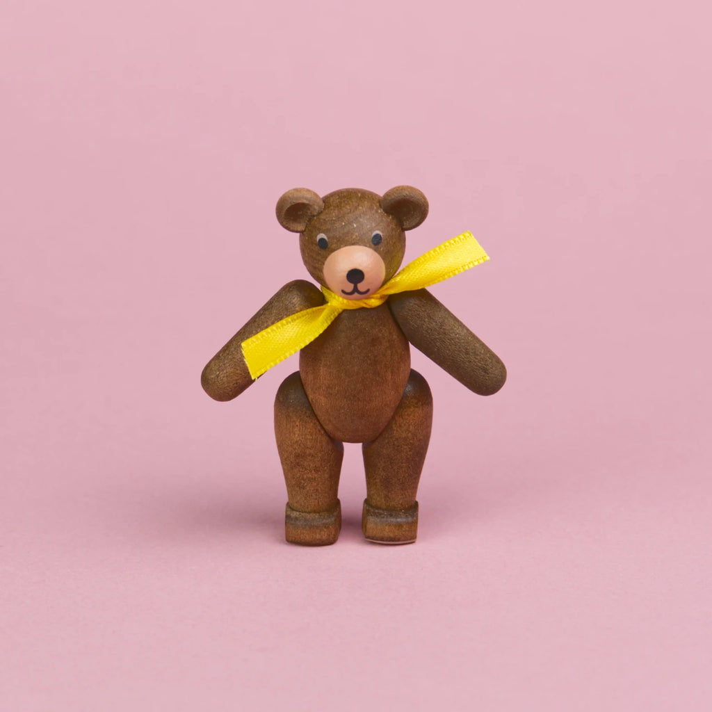 ein kleiner Teddybär aus dunklem Holz mit gelber Schleife um den Hals, Beine und Arme sind beweglich , er ist in einer stehenden Position zu sehen, er hat eine hellere Schnauze und seine Augen und Nase sind schwarz , sein Mund lächelt