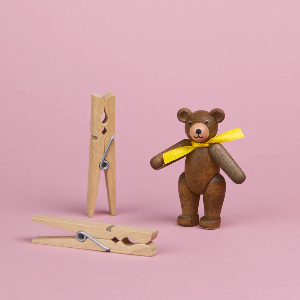 ein kleiner Teddybär aus dunklem Holz mit einer gelber Schleife um den Hals steht neben 2 Wäscheklammern, die beiden Objekte sind annähernd gleich groß, seine Augen und Nase sind schwarz und er hat eine hellere Schnauze