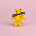 ein kleiner Teddybär aus gelb bemaltem Holz mit grüner Schleife um den Hals, Beine und Arme sind beweglich und er ist in einer stehenden Position zu sehen, seine Augen und Nase sind schwarz und sein Mund lächelt