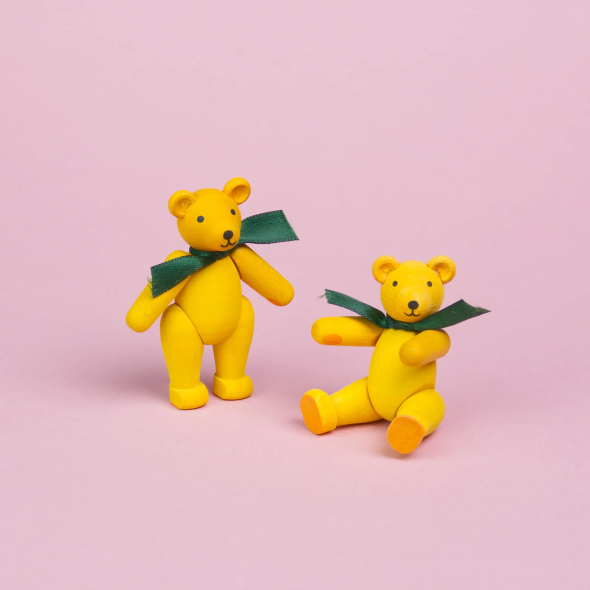 Zwei kleine Teddybären aus gelb bemaltem Holz mit grünen Schleifen um den Hals, Beine und Arme sind beweglich , einer ist in einer stehenden Position zu sehen der andere sitztund zeigt seine orangenen Pfoten, beide haben schwarze Augen und Nasen