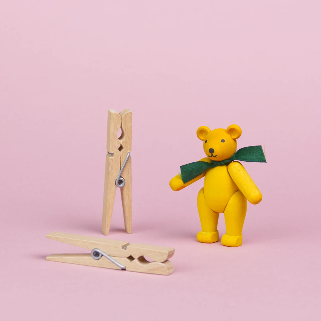 ein kleiner Teddybär aus gelb bemaltem Holz mit einer grüner Schleife um den Hals steht neben 2 Wäscheklammern, die beiden Objekte sind annähernd gleich groß, seine Augen und Nase sind schwarz und sein Mund lächelt