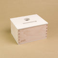 Eine rechteckige Holzbox von Grapat steht seitlich eingedreht in der Mitte, sodass drei Seiten der Box zu sehen sind. Sie hat einen Deckel in dem eine längliche und eine Runde Einkerbung sind, sodass Dinge in dieser Form in die Box geworfen werden können.