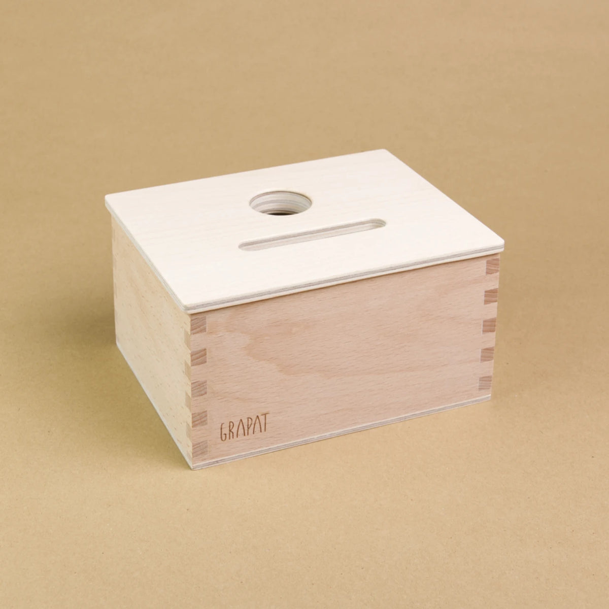 Eine rechteckige Holzbox von Grapat steht seitlich eingedreht in der Mitte, sodass drei Seiten der Box zu sehen sind. Sie hat einen Deckel in dem eine längliche und eine Runde Einkerbung sind, sodass Dinge in dieser Form in die Box geworfen werden können.