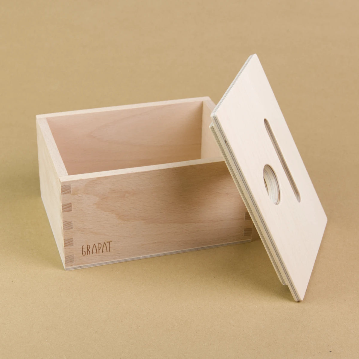 Es ist eine rechteckige Holzbox von Grapat zu sehen. Sie hat einen Deckel in dem eine längliche und eine Runde Einkerbung sind, sodass Dinge in dieser Form in die Box geworfen werden können. Der Deckel ist seitliche an die vordere, rechte Ecke der Box gelehnt.