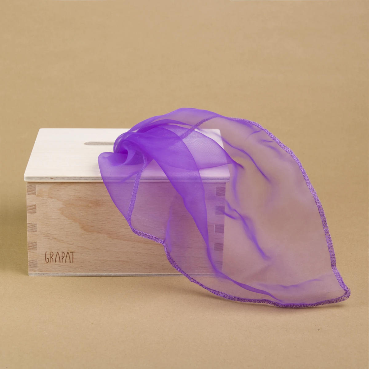 Eine rechteckige Permanente Box aus Holz steht gerade im linken Teil des Bildes. Nach rechts erstreckt sich ein violett färbendes, transparentes Tuch, das aus einer Öffnung des Deckels der Box kommt.