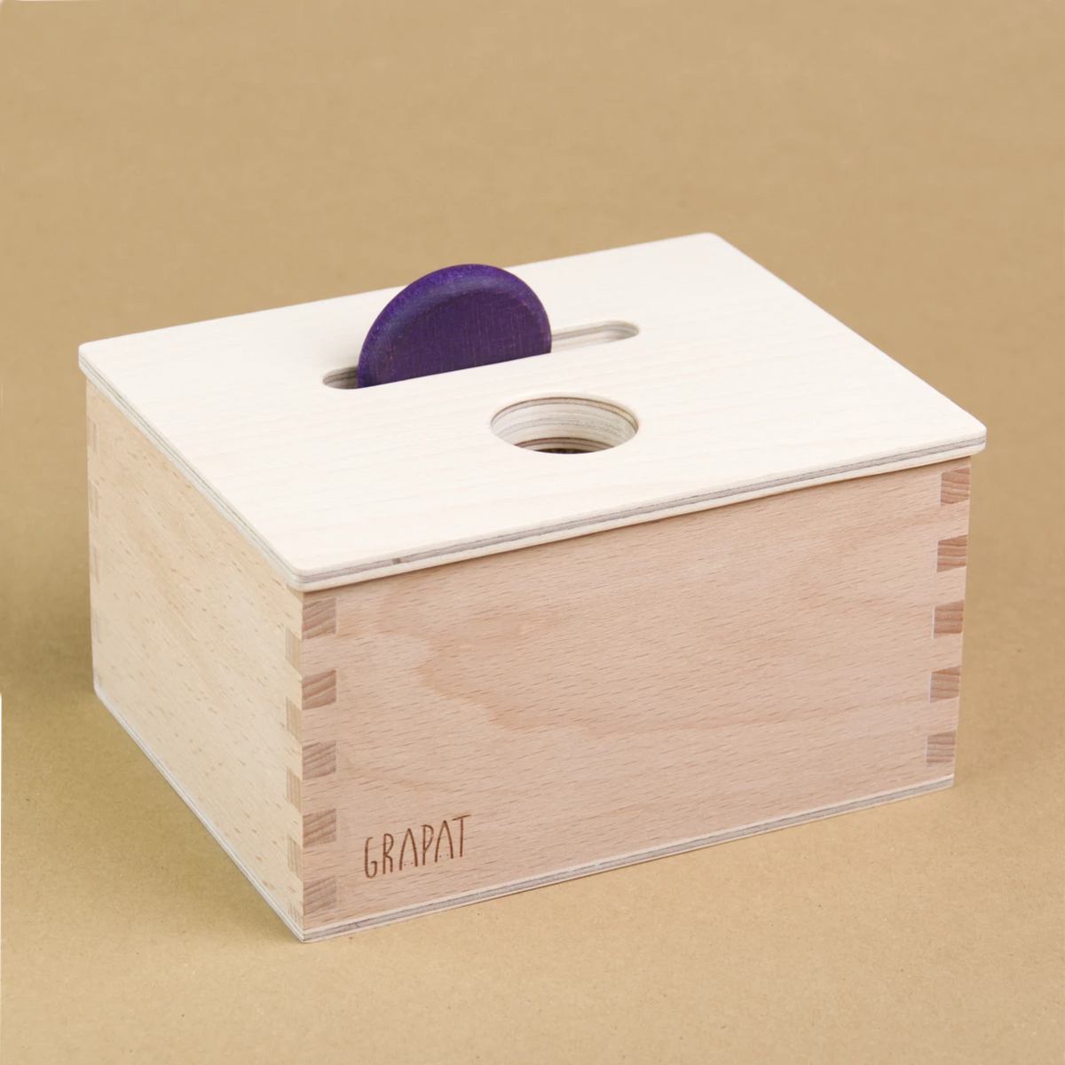 Eine rechteckige Holzbox von Grapat steht seitlich eingedreht in der Mitte, sodass drei Seiten der Box zu sehen sind. Sie hat einen Deckel in dem eine längliche und eine Runde Einkerbung sind. In der länglichen Einkerbung steckt eine violette Holzmünze.