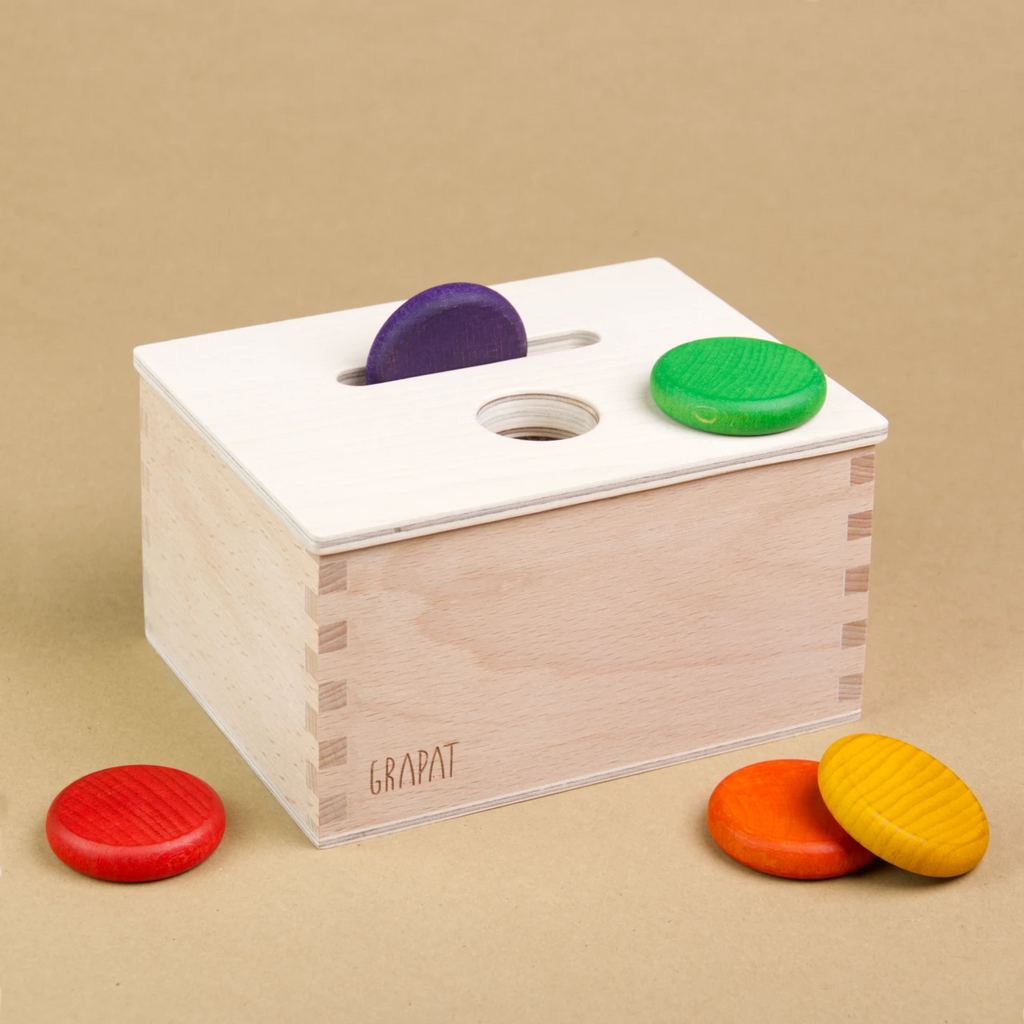 Eine rechteckige Permanence Box von Grapat aus Holz steht seitlich eingedreht da, sodass drei Seiten der Box zu sehen sind. Sie hat einen Deckel in dem eine längliche und eine Runde Einkerbung zu sehen sind. In der länglichen Einkerbung steckt eine violette Holzmünze. Auf dem Deckel liegt außerdem eine grüne Holzmünze und vor und neben der Holzbox sind auch eine rote, eine gelbe und orangene Holzmünze zu sehen.
