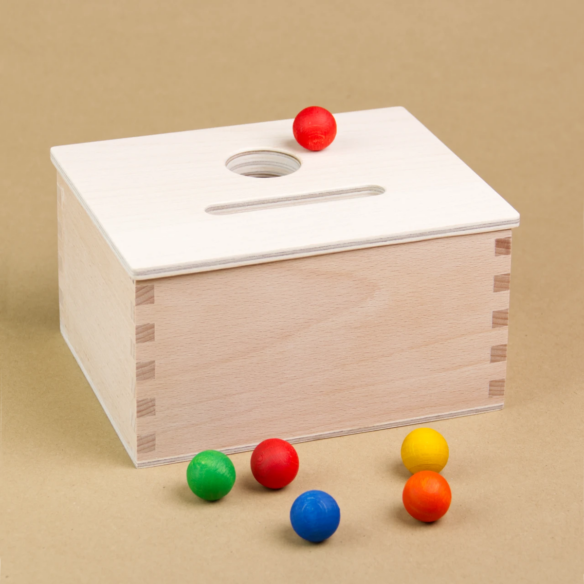 Eine rechteckige Permanence Box von Grapat aus Holz steht seitlich eingedreht da, sodass drei Seiten der Box zu sehen sind. Sie hat einen Deckel in dem eine längliche und eine Runde Einkerbung zu sehen sind. Neben der runden Einkerbung liegt eine rote Holzmurmel. Vor der Holzbox sind auch eine weitere rote, eine gelbe, orangene, grüne und blaue Holzmurmel zu sehen.