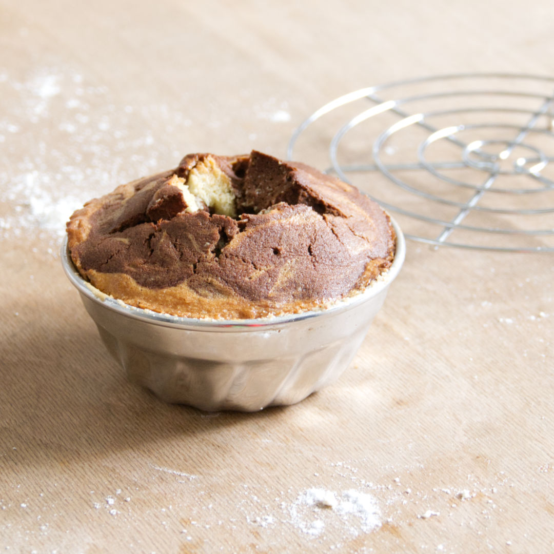 Komposition aus einer mit gebackenem Kuchenteig gefüllte Gugelhupfform aus Alu und Drahtuntersetzer von Glückskäfer.