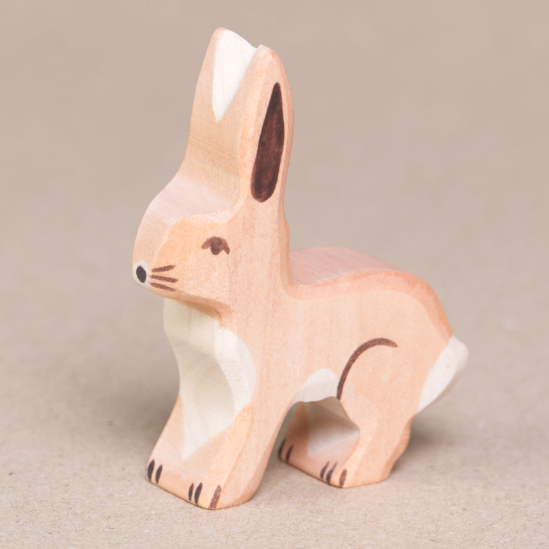 Ein Hase mit nach oben hin stehenden Ohren von Holztiger, hat hellbraunes Fell, einen weißen Schwanz sowie Bauchfell. Es schaut nach links.