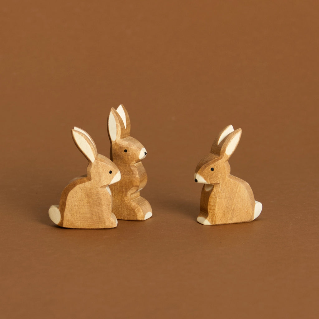 Komposition aus Hase sitzend, rechts im Bild und Hase mit Ohren hoch hinten links und vorne links nochmals Hase sitzend. Alle haben ihre Ohren nach oben gespitzt und schauen zur Bildmitte hin.