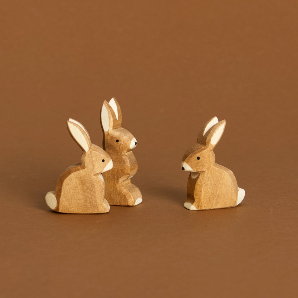 Komposition aus Hase sitzend, rechts im Bild und Hase mit Ohren hoch hinten links und vorne links nochmals Hase sitzend. Alle haben ihre Ohren nach oben gespitzt und schauen zur Bildmitte hin.