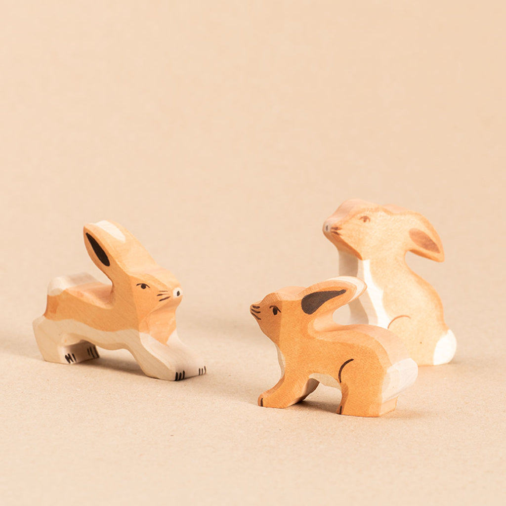 Drei hellbraune Holzhasen mit weißem Bauchfell und weißer Blume. Der linke Hase laufend mit nach oben hin gespitzten Ohren, schaut nach vorne rechts. Der in der Bildmitte stehende Hase schaut mit waagerechten Ohren nach hinten links und der weiter im hintergrund im rechten Bildteil sitzende Hase mit nach unten hin zeigenden Ohren schaut nach links oben.