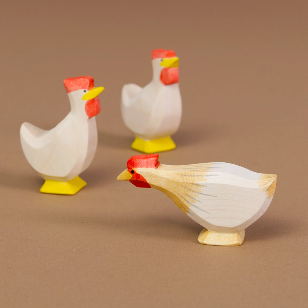 Komposition aus Huhn ocker lang und zwei Hühner in weiß. Im Vordergrund befindet sich das nach links schauende Huhn ocker lang und im Hintergrund die anderen Hühner in weiß. Alle haben einen auffällig roten Haarkamm.