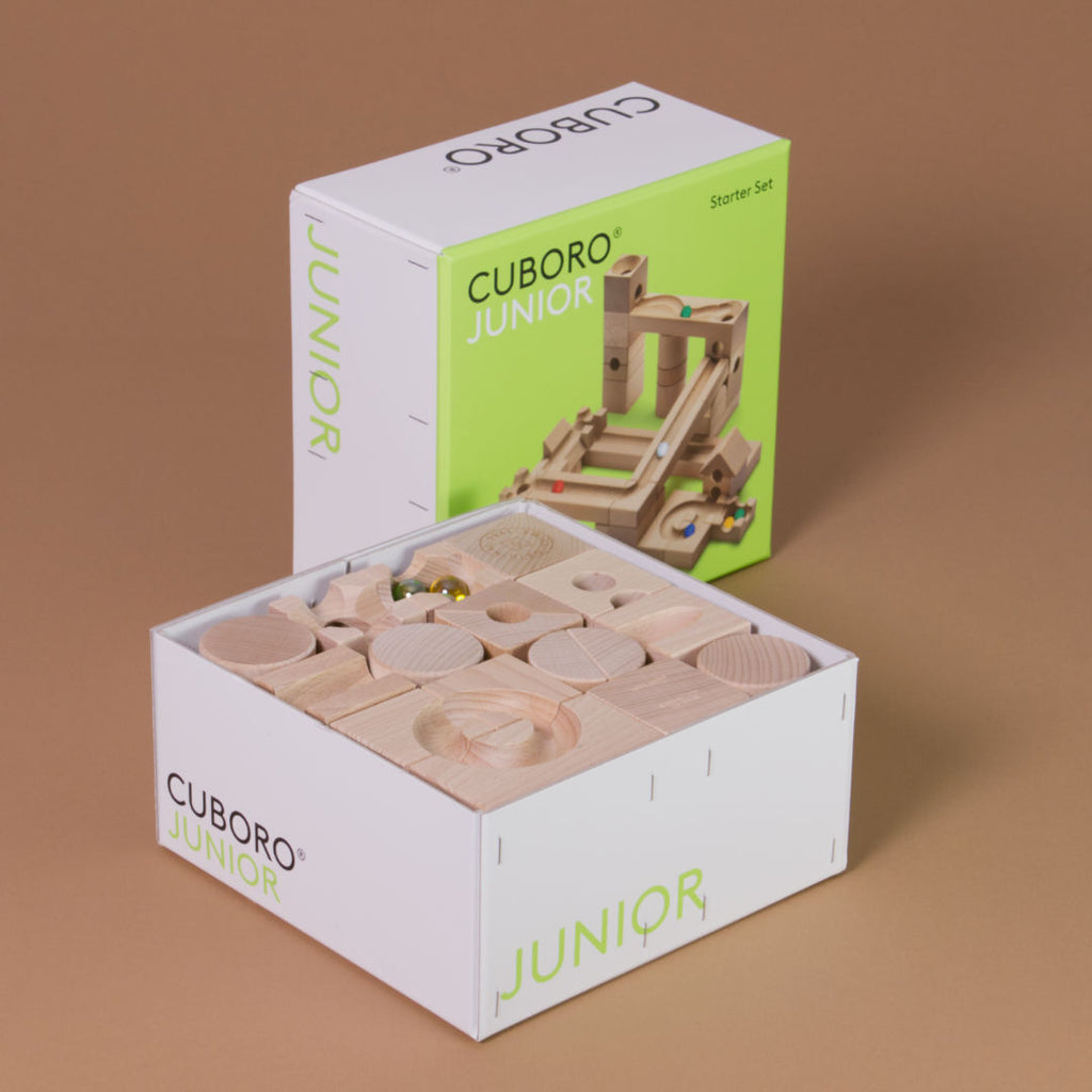Das Junior Starter Set von Cuboro aus Buche gefertigt liegt im Produktkarton und geöffnetem Kartondeckel da, welcher im Hintergrund senkrecht aufgestellte ist.