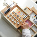 Spielzeugkasse aus Holz und in Creme farben, runden Knöpfen und Spielgeld im Geldfach. Daneben liegen Holz-EC-Karten-Lesegerät, Holz-EC-Karte und Holz-Warenscanner. Kinderhand nimmt Papiergeld aus Schublade.