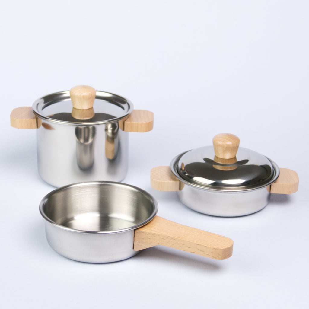 Es ist in Kochset von der Marke Goki zu sehen. Dabei handelt es sich um eine Pfanne mit Holzgriff vorne, eine höheren Topf mit Deckel und Holzgriffen hinten links, sowie einem kleineren Topf mit Deckel und ebenfalls Holzgriffen hinten rechts.