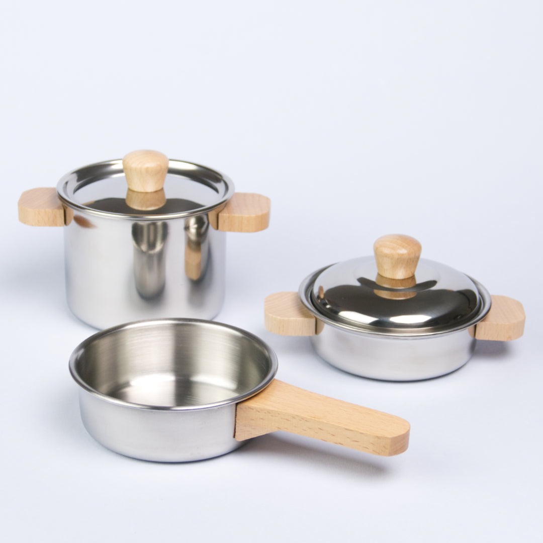 Es ist in Kochset von der Marke Goki zu sehen. Dabei handelt es sich um eine Pfanne mit Holzgriff vorne, eine höheren Topf mit Deckel und Holzgriffen hinten links, sowie einem kleineren Topf mit Deckel und ebenfalls Holzgriffen hinten rechts.