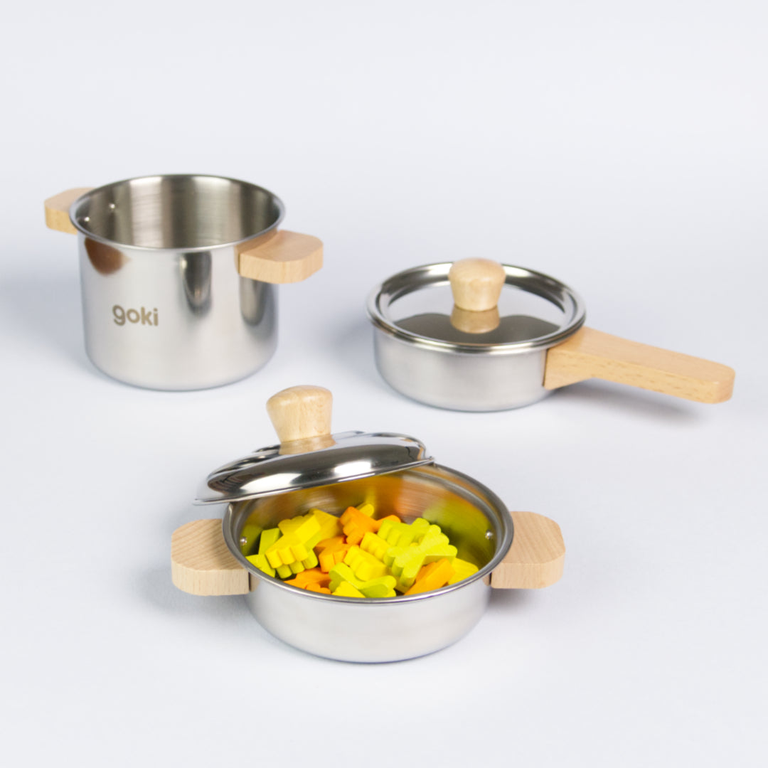 Es ist in Kochset von der Marke goki zu sehen. Abgebildet ist eine kleinerer Topf mit Deckel und gelben, grünen und orangenen Farfalle darin, vorne, ein höherer Topf mit Deckel und Holzgriffen hinten links, sowie einer Pfanne mit ebenfalls Holzgriffen hinten rechts.