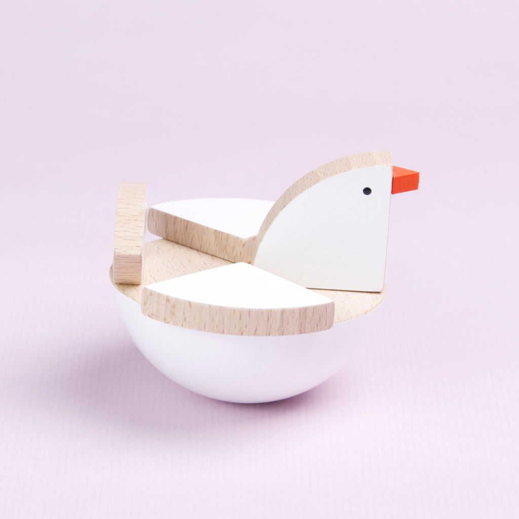 Es ist ein Schaukelvogel aus Holz von der Seite zu sehen, der nach rechts schaut. Der untere Teil des Vogels besteht aus einer großen Halbkugel, die weiß bemalt ist und an der Oberseite holzfarben ist. Darauf sitzen mehrere dreiecksförmige Holzteile, die ebenfalls weiß bemalt sind, bis auf die Kanten. Der Schnabel ist orange eingefärbt.