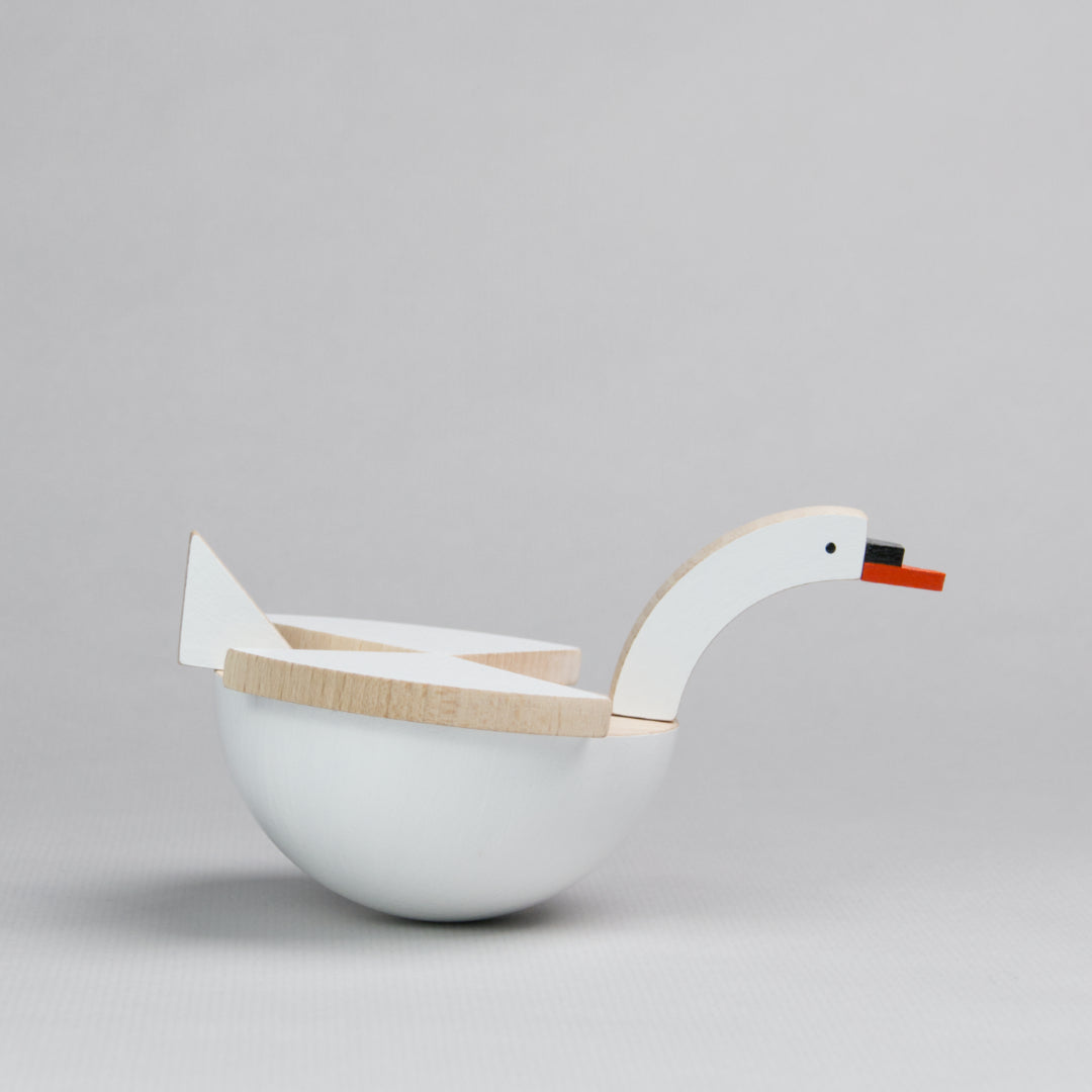 Es ist ein beweglicher Schwan zu sehen, der von der Seite zu sehen ist, nach rechts schaut und dabei leicht nach vorne kippt. Sein Körper besteht aus einer Halbkugel, die weiß bemalt ist, darauf sitzen drei Dreiecke, die die Flügel und die Schwanzfedern darstellen. Vorne befindet sich ein langer, weißer Hals der mit einem rot-schwarzen Schnabel endet.