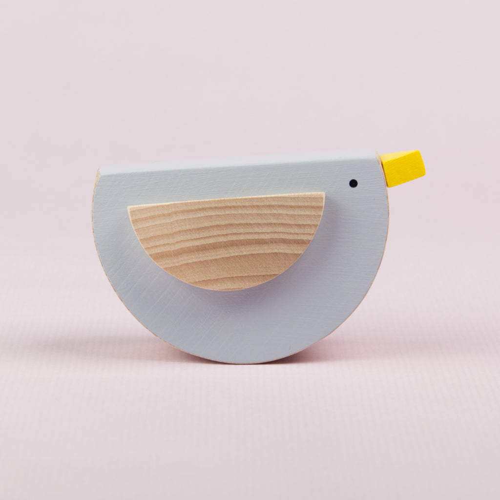 Es ist ein schwingender Vogel aus Buchenholz von der Seite zu sehen. Sein Bauch ist grau bemalt. Ein Flügel ist zu sehen, welcher Holzfarben ist und der Schnabel ist gelb bemalt.