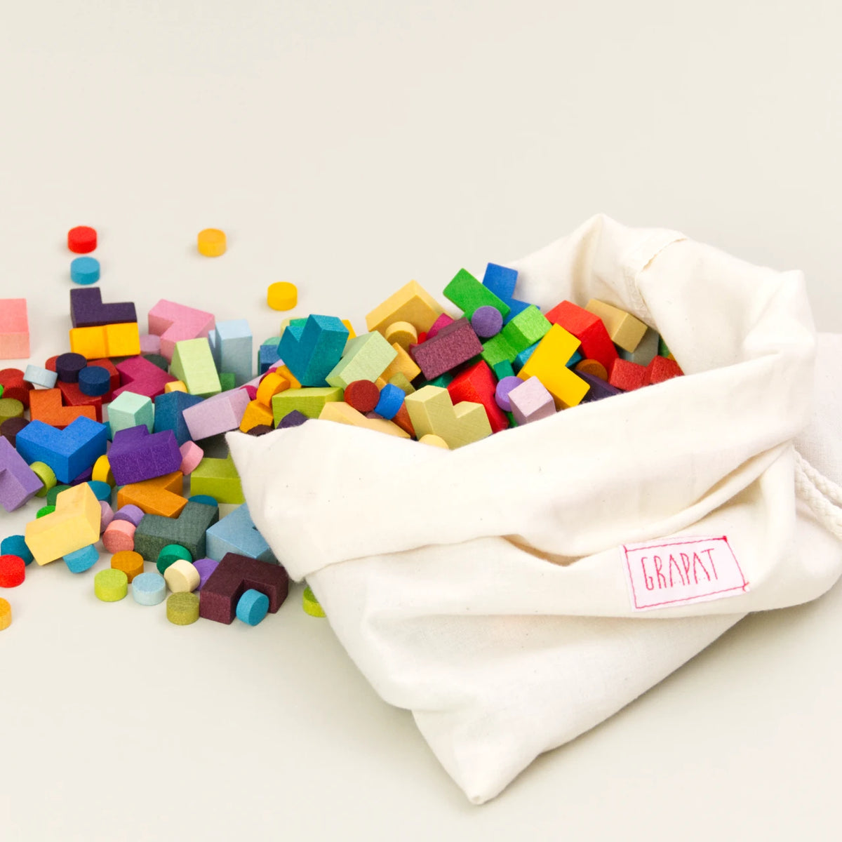 Grapat Mis Match and Bloom, Mosaik Elemente, 400 Teile, 36 Farbnuancen – bunte Holzbausteine in verschiedenen Formen und Farben, teilweise zylindrisch und eckig, ausgeschüttet aus einem weißen Stoffbeutel mit rotem Grapat-Logo, Darstellung des gesamten Spielmaterials
