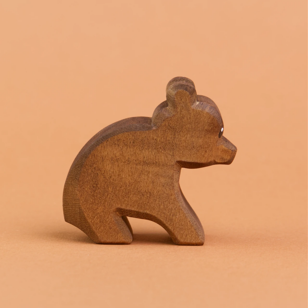 Ein kleiner, sitzender Bär aus Holz ist in der Seitenansicht zu sehen und schaut nach rechts. Er hat dunkelbraunes Fell und runde, hochstehende Ohren. Er ist von der Marke Ostheimer.