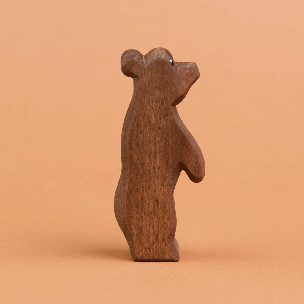 Ein kleiner Bär aus Holz ist stehend in der Seitenansicht zu sehen und schaut nach rechts, oben. Er hat dunkelbraunes Fell und große, runde Ohren. Er ist von der Marke Ostheimer.