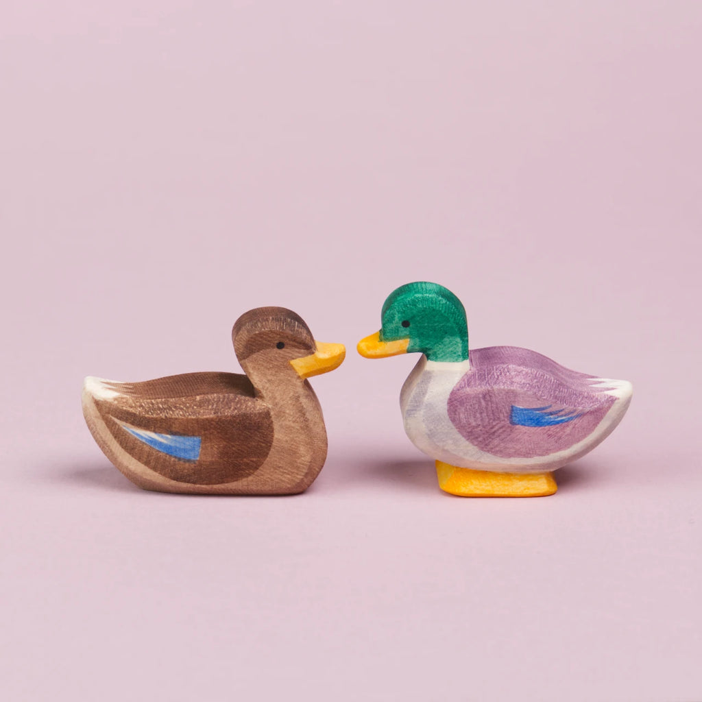 Ein Erpel rechts und eine Ente links aus Holz sitzen sich gegenüber und sind in der Seitenansicht zu sehen. Sie schauen sich an und berühren sich fast mit ihrem gelben Schnabel.