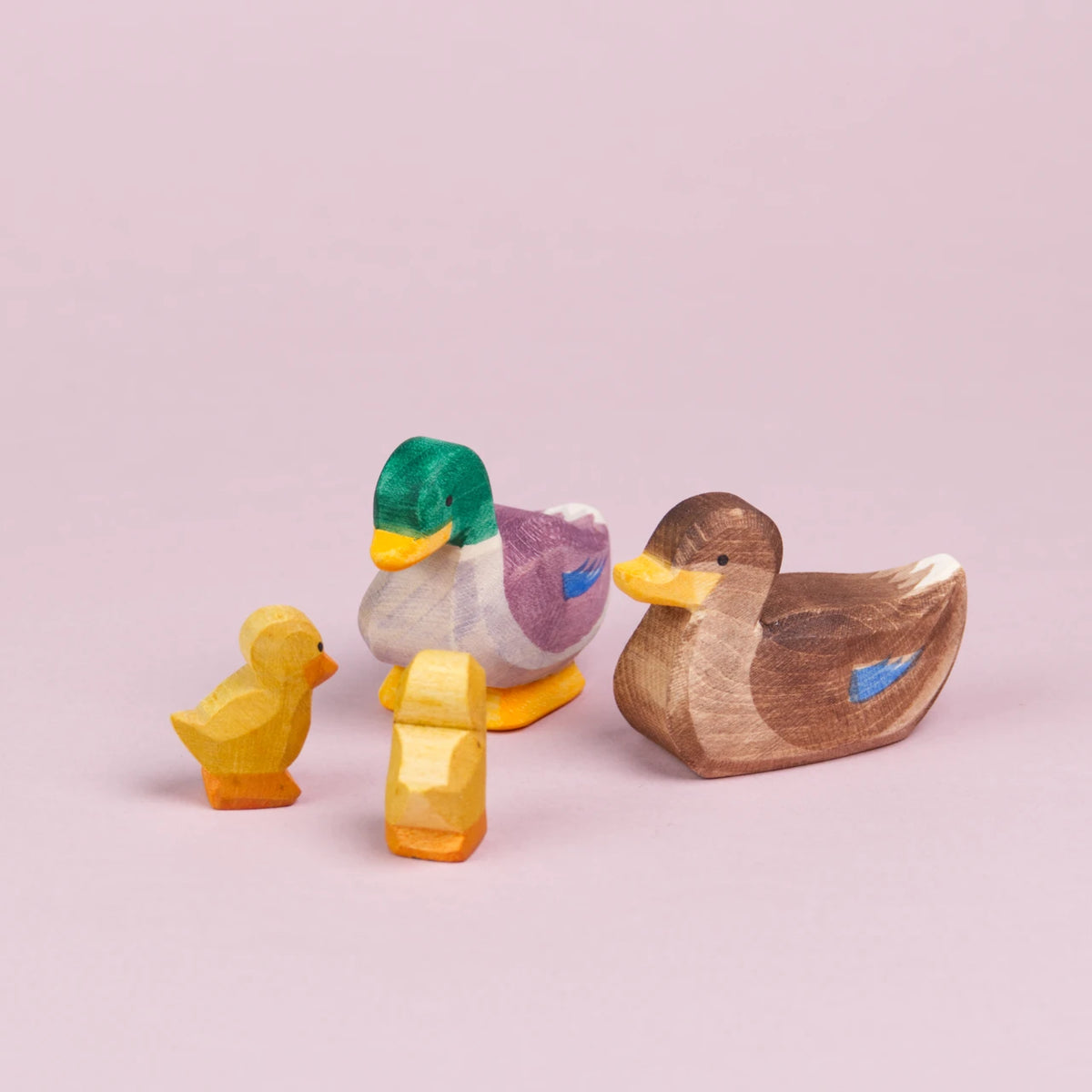 Es ist eine Entenfamilie aus Holz von der Mark Ostheimer zu sehen. Hinten in der Mitte sitzt ein Erpel mit grünen, lilafarbenen und weißen Federn, welcher nach vorne links unten auf zwei kleine Entenküken schaut. Rechts neben ihm sitzt eine Ente mit hellbraunen und dunkelbraunen Federn, die wie bei dem Erpel an den Flügeln blau bemalt sind. Die Ente schaut ebenfalls nach vorne links zu den Küken herüber.