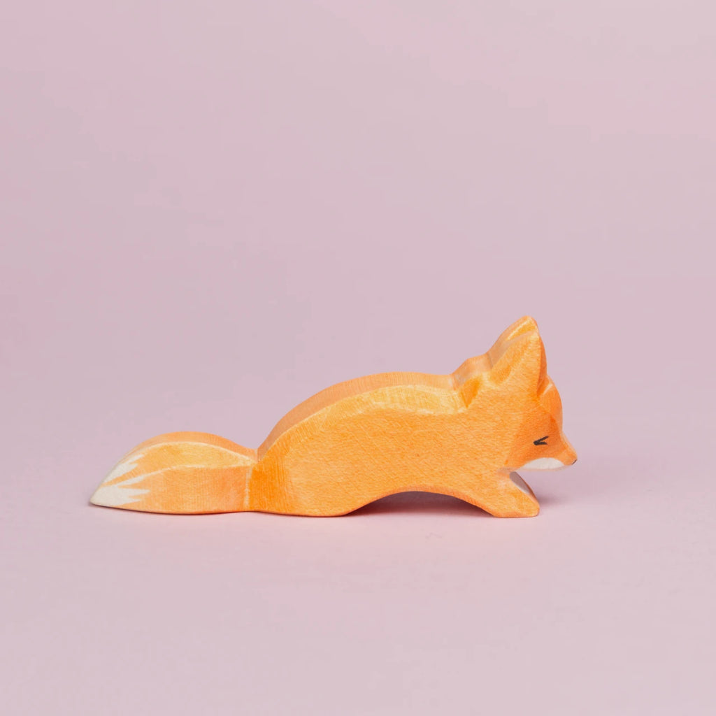 Es ist ein kleiner, schleichender Fuchs aus Holz von der Seite zu sehen, der nach rechts schaut. Sein Fell ist orangefarben und an seinem Maul und dem Ende seines Schwanzes ist es weiß. Er schaut nach unten und ist von der Marke Ostheimer.