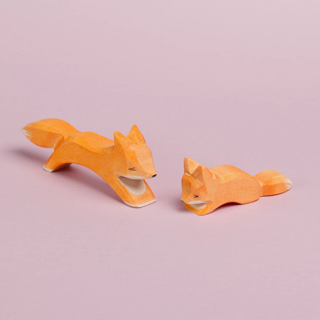 Links ist in großer, laufender Fuchs zu sehen, der nach vorne rechts gerichtet ist und einen kleinen, schleichenden Fuchs links von ihm anschaut, der nach vorne links gerichtet ist und nach unten schaut.