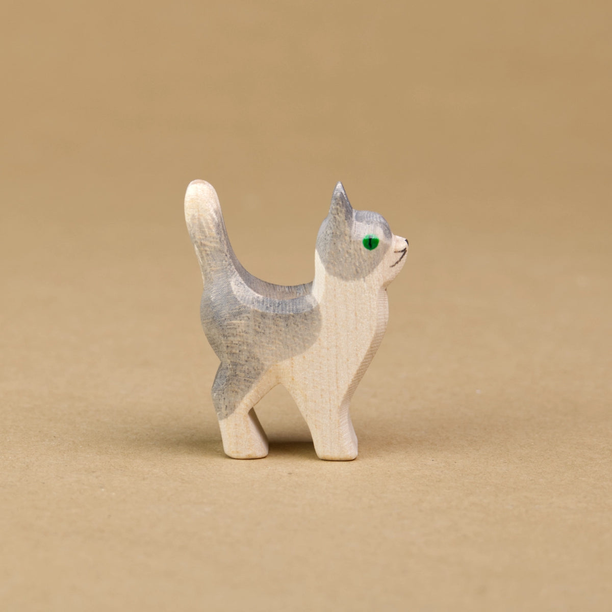 Eine kleine Katze aus Holz von der Marke Ostheimer ist von der Seitenansicht zu sehen und hält ihren Kopf, sowie auch den Schwanz nach oben. Der Großteil ihres Felles ist weiß gefärbt und der obere Part des Kopfes mit den Ohren, sowie der Rücken über einen Teil der Hinterbeine bis zu den Schwanz ist dunkelgrau eingefärbt. Die Augen der kleinen Katze sind dunkelgrün.