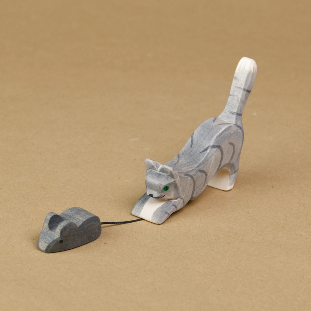 Eine springende Katze ist seitlich eingedreht zu sehen, wie sie eine Maus fängt. Sie streckt den Kopf und den Oberkörper nach unten dicht an den Boden und hält mit den Vorderpfoten den Schwanz einer Maus fest. Ihr Hinterteil streckt die Katze nach oben. Die Maus, sowie die Katze schauen nach vorne links und sind von der Marke Ostheimer.