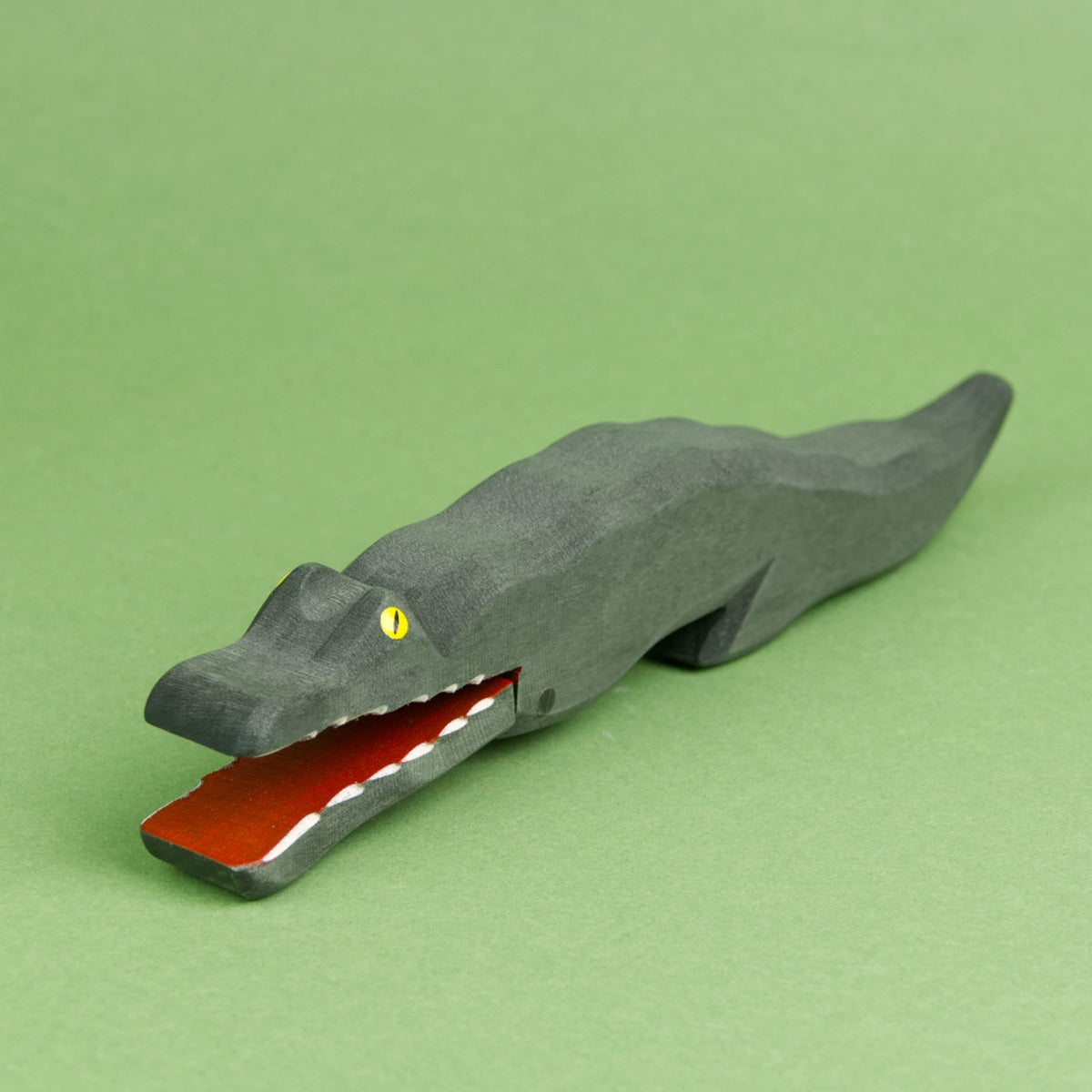 Ein Krokodil aus Holz ist schräg von der Seite zu sehen. Der Rücken hat eine leicht wellige Oberfläche und es hat das Maul weit geöffnet, wodurch die weißen, langen Zähne zu sehen sind. Innen ist das Maul des Krokodils rot bemalt.
