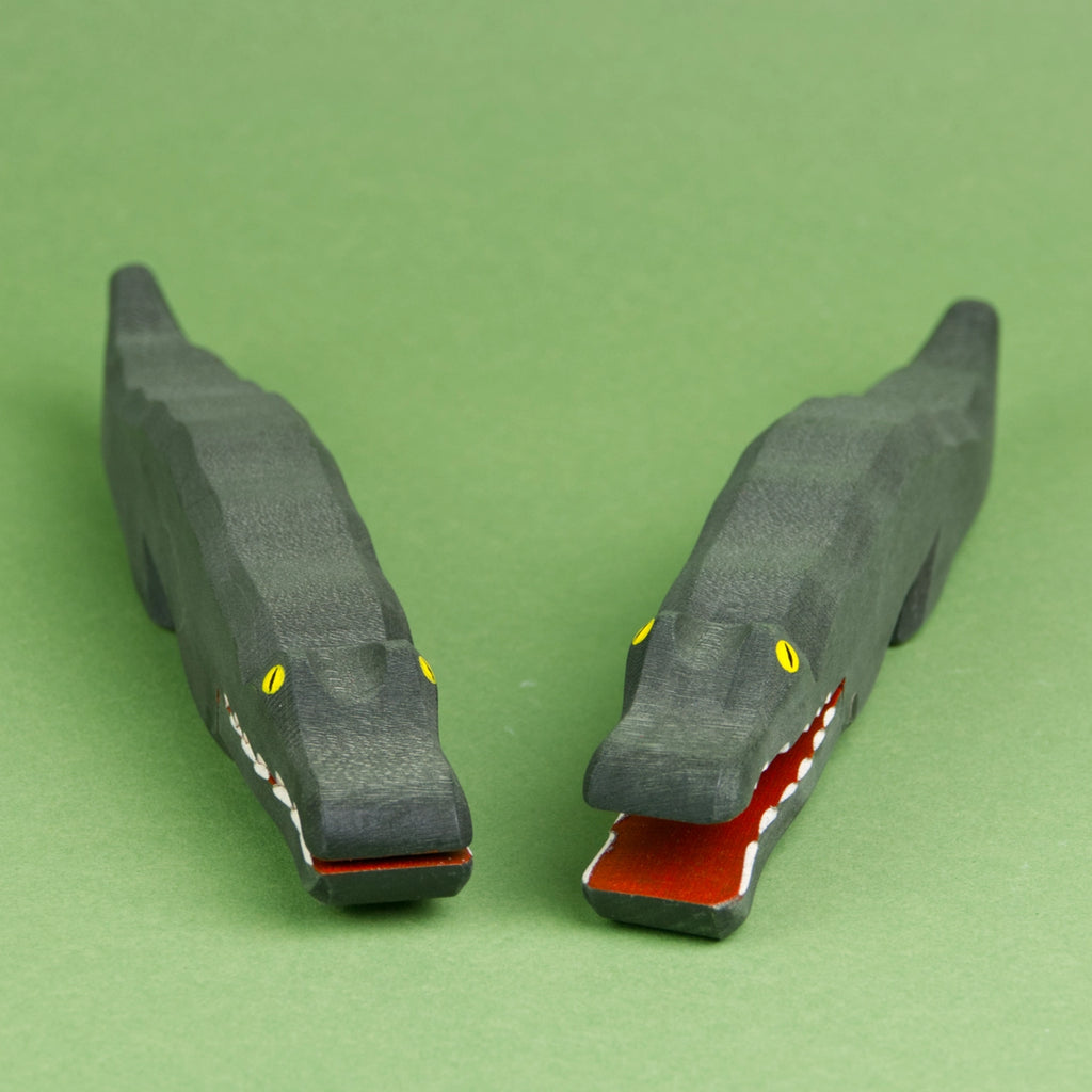 Zwei Krokodile aus Holz sind schräg nebeneinander zu sehen. Das Hinterteil des rechten Krokodils ist leicht nach rechts außen gedreht, das des linken nach links außen, sodass die Köpfe näher zusammen sind. Das rechte Krokodil hat sein Maul weit geöffnet, das linke hat es geschlossen.