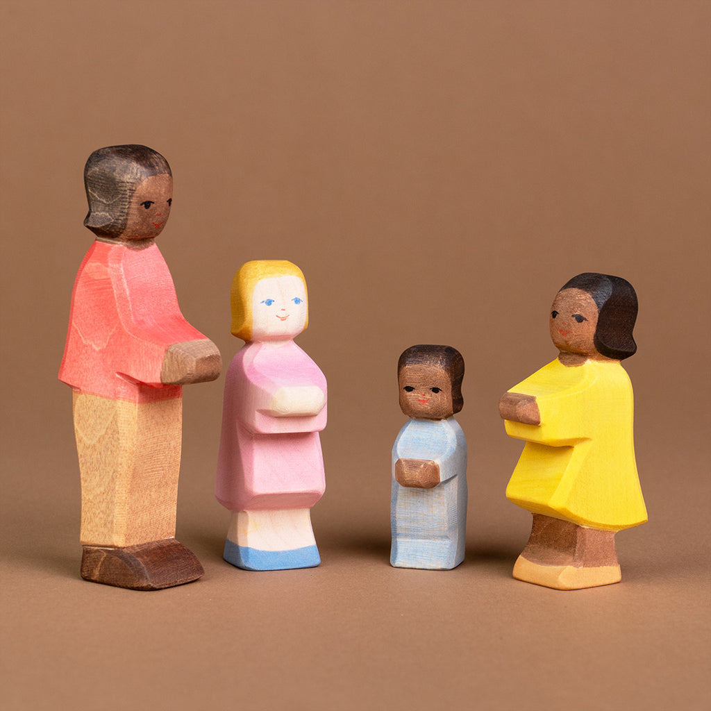 Es ist eine Gruppe von Kindern zu sehen. Ganz links steht ein Sohn mit dunkler Hautfarbe seitlich eingedreht und schaut nach vorne rechts. Daneben stehen eine Tochter mit heller Hautfarbe und ein Wickelkind mit dunkler Hautfarbe, die beide nach vorne schauen. Rechts von ihnen steht eine Tochter mit dunkler Hautfarbe, die nach vorne links schaut.